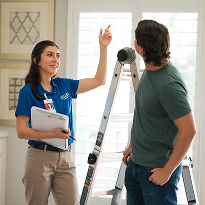 Female Aire Serv tech standing in front of ladder pointing to ceiling.